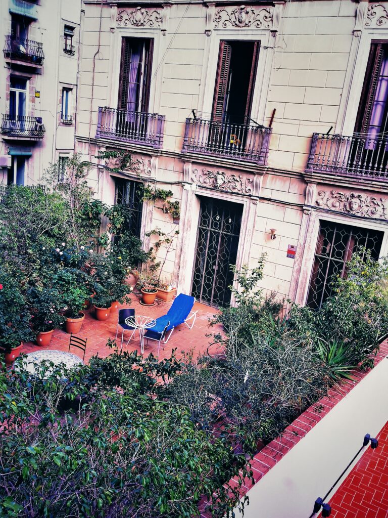 Dies ist ein Farbfoto eines Hinterhes eines alten Hauses in Barcelona. Man sieht eine Terasse, die zu 2/3 vollgestellt ist mit Grün- und Blühpflanzen, eine Liege mit blauem Bezug vor einem kleinen runden Tisch, an dem noch ein Metallstuhl steht. Es wirkt gemütlich.